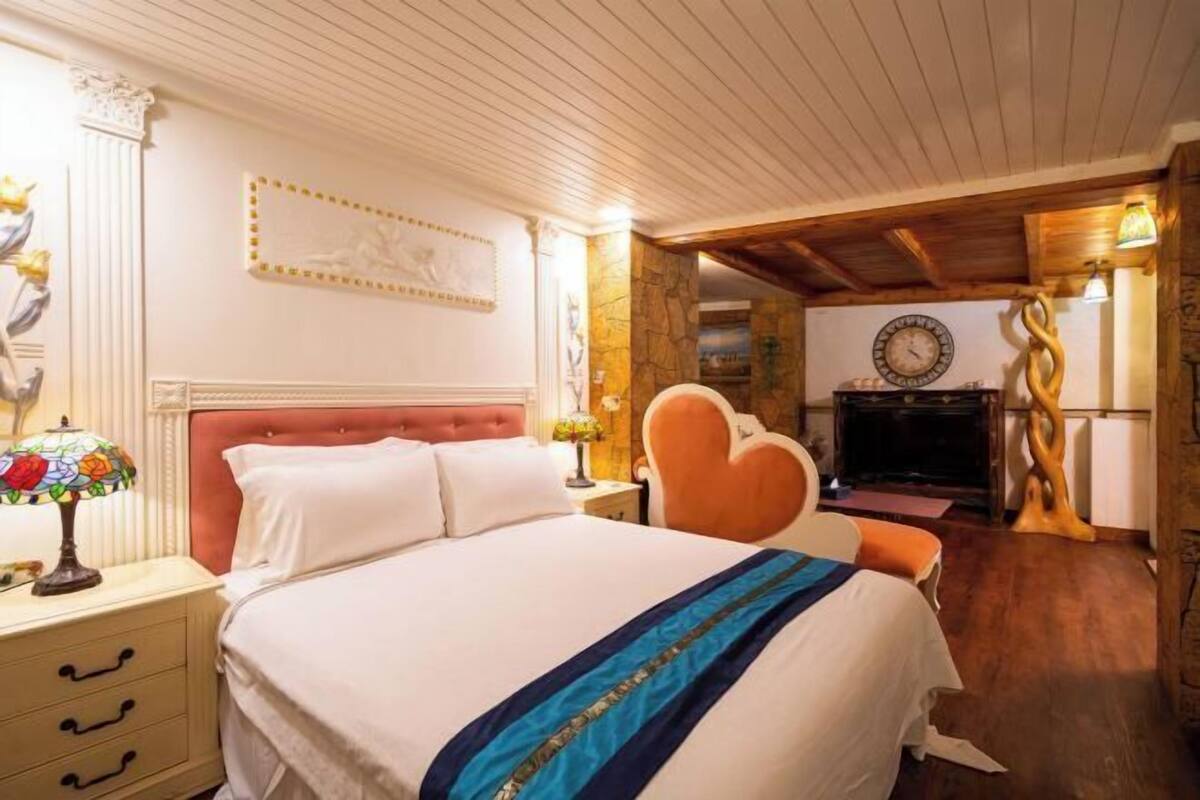 Een slaapkamer met een bed, een nachtkastje, een hartvormige fauteuil, een open haard en een klok aan de muur.