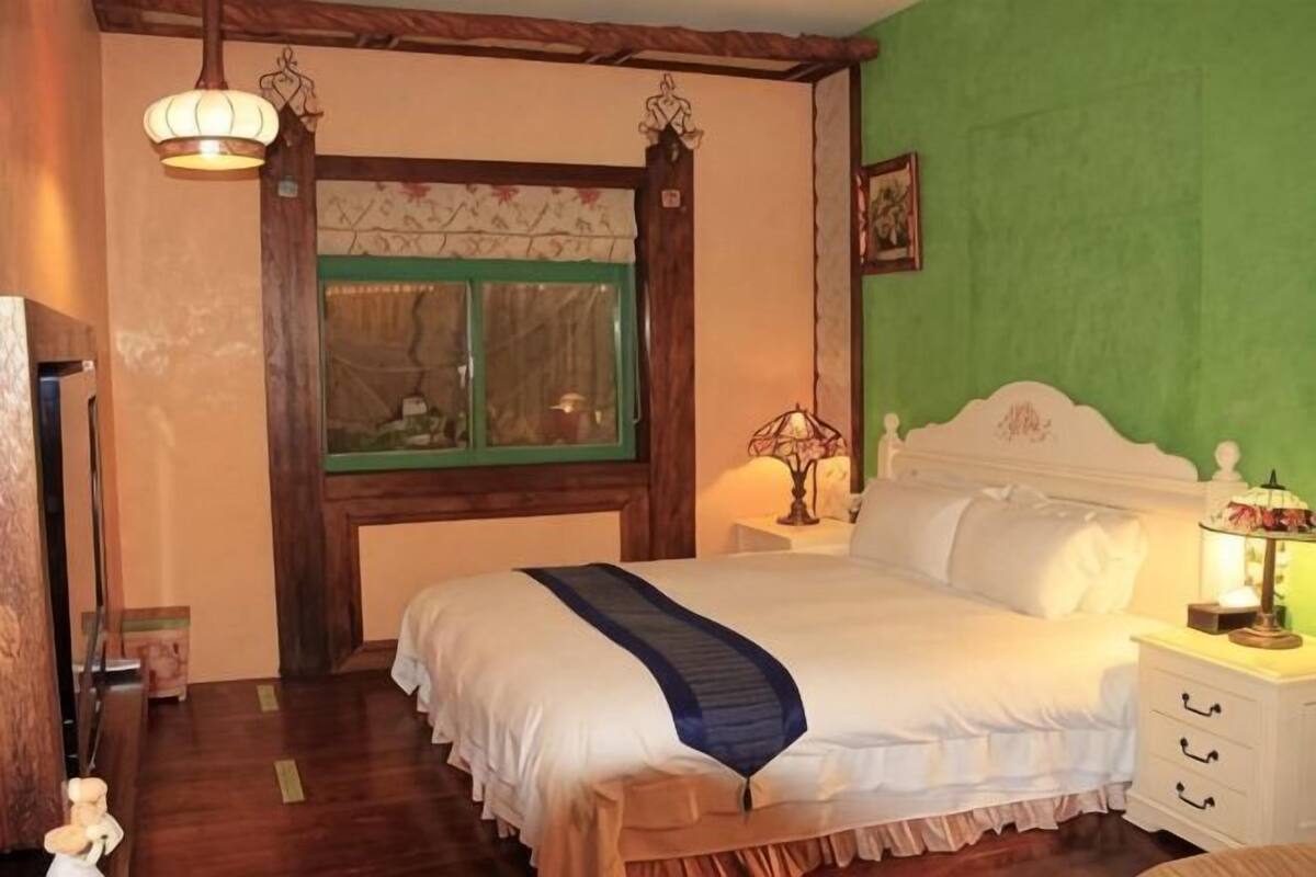 Een slaapkamer met een bed, nachtkastje, lamp, raam en houten vloer.