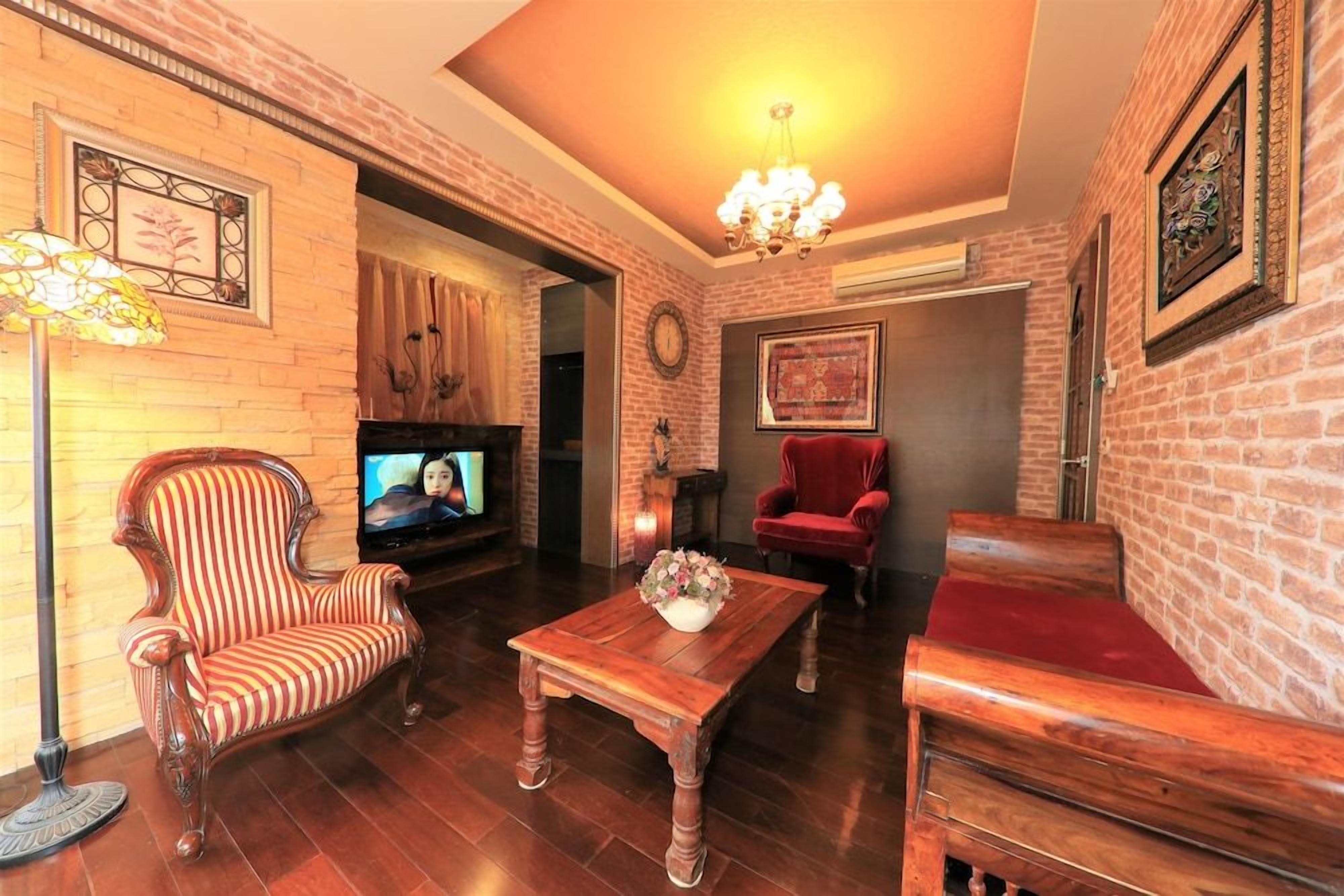 Sala de estar com parede de tijolos, televisão, poltrona vermelha, mesa de centro de madeira e um candelabro.