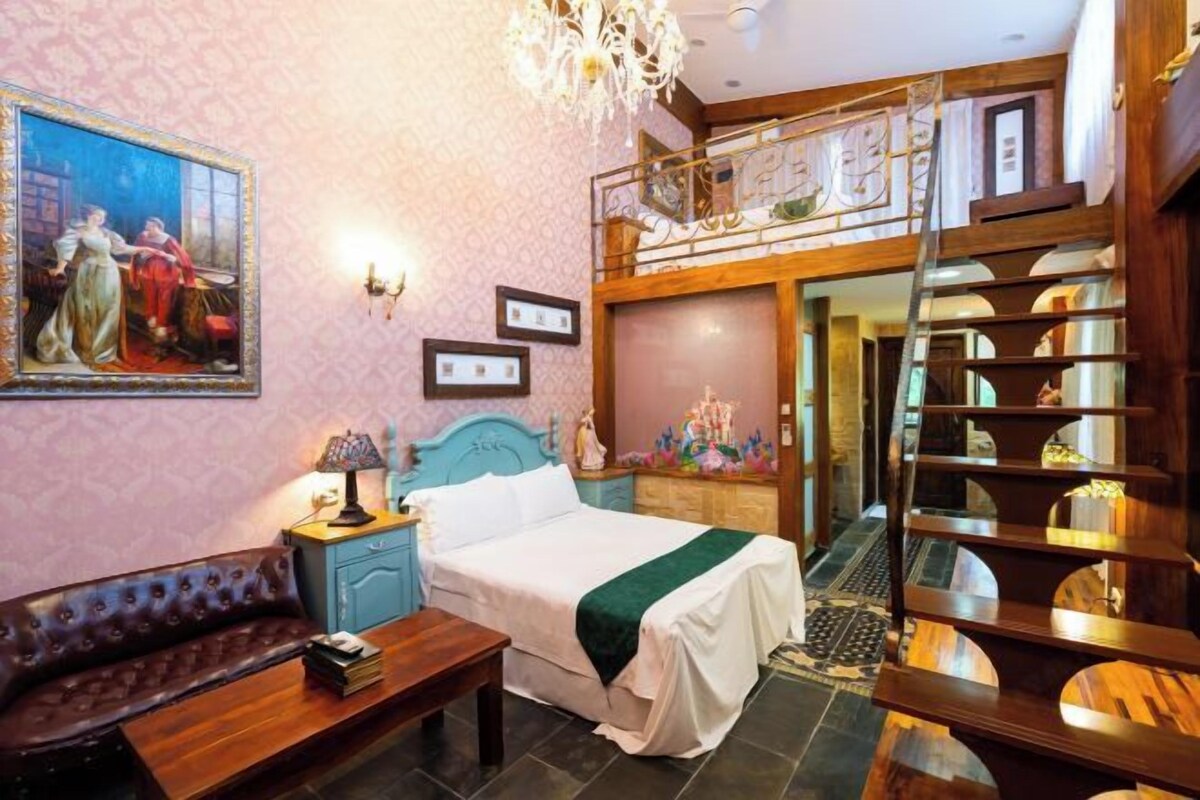 Ein Schlafzimmer mit einem Bett, einem Nachttisch, einer Kommode, einer Bank, einem Gemälde und einer Treppe, die zu einem Schlafdach führt.