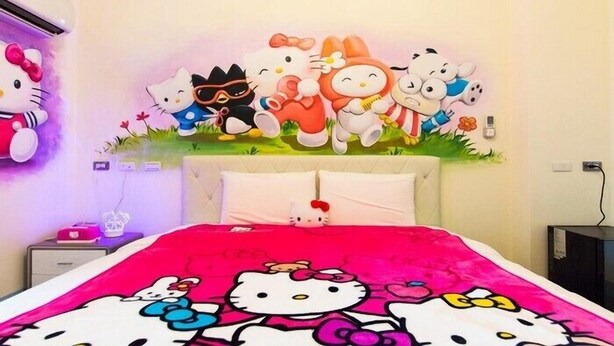 Un dormitorio con cama decorada con diseño de Hello Kitty, un mural en la pared con personajes de Hello Kitty y una mesita de noche con una lámpara de Hello Kitty.
