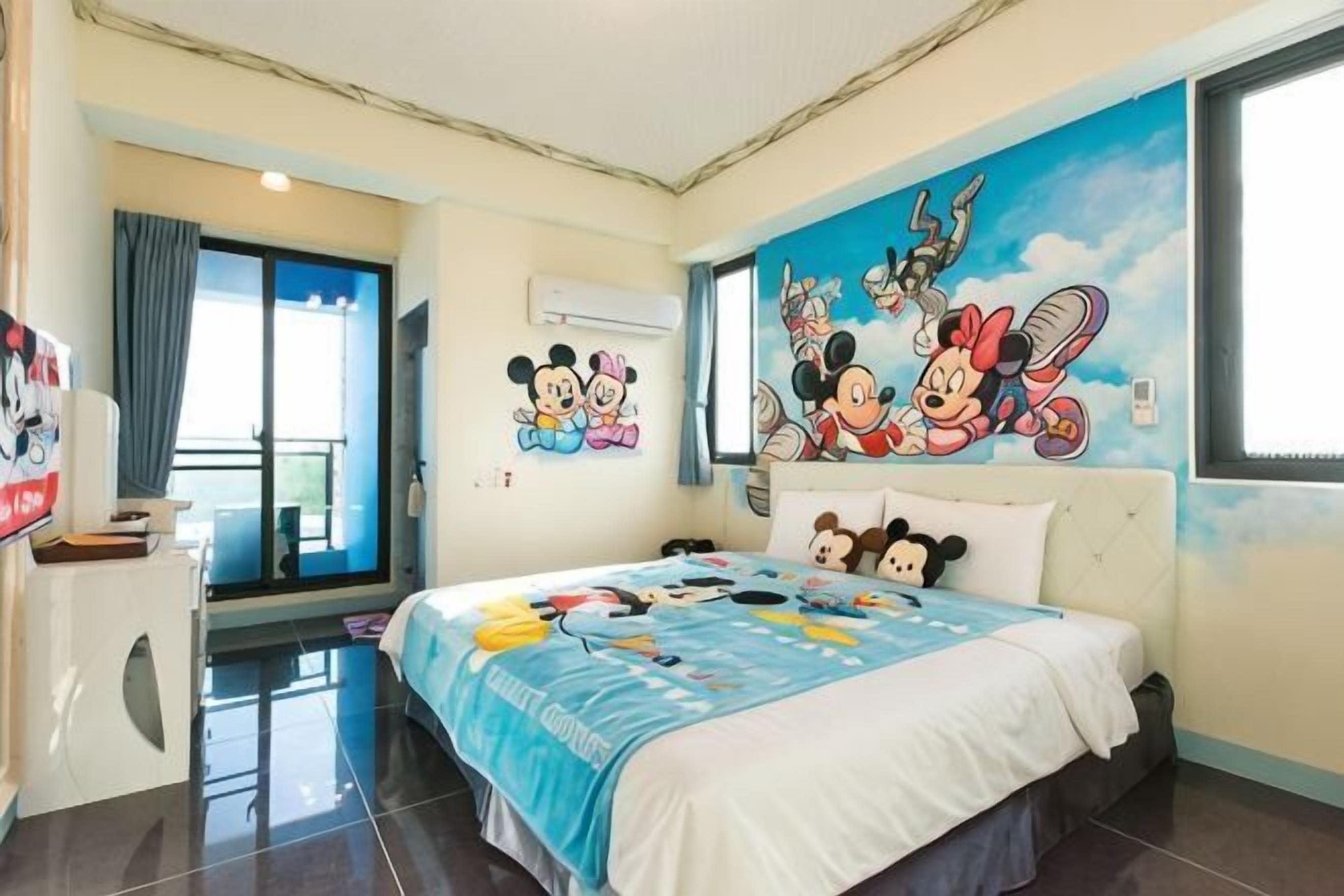 A hotel room with a bed, a desk, a window, and a mural of cartoon characters.