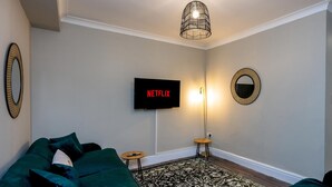 Una sala de estar con un sofá verde, un televisor de pantalla plana montado en la pared, una lámpara de pie y una alfombra estampada.