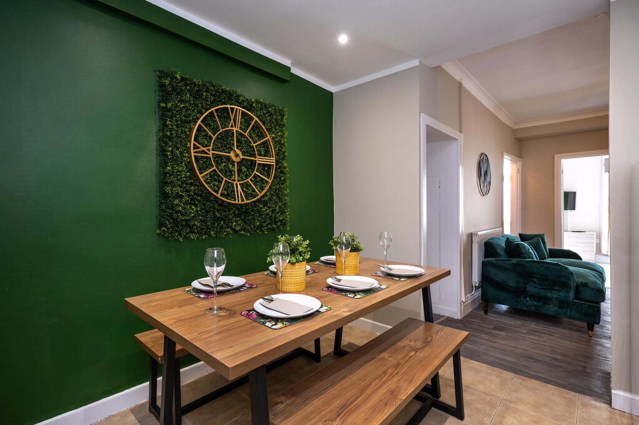 Un comedor con una mesa de madera dispuesta para cuatro, una pared verde con un reloj y un sofá verde al fondo.