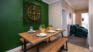 A dining area with a wooden table set for four, a green wall with a clock, and a green sofa in the background. - Comfortable 4-bed Home With Free Parking Wales (Mountain Ash)