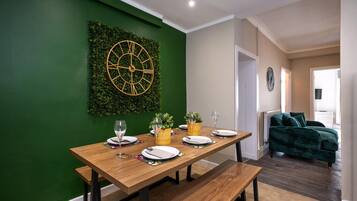 Un comedor con una mesa de madera dispuesta para cuatro, una pared verde con un reloj y un sofá verde al fondo.