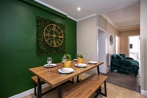 A dining area with a wooden table set for four, a green wall with a clock, and a green sofa in the background. - Comfortable 4-bed Home With Free Parking Wales (Mountain Ash)