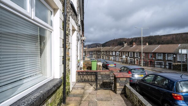 A residential street with terraced houses, parked cars, and a closed shop window. - Comfortable 4-bed Home With Free Parking Wales (Mountain Ash)
