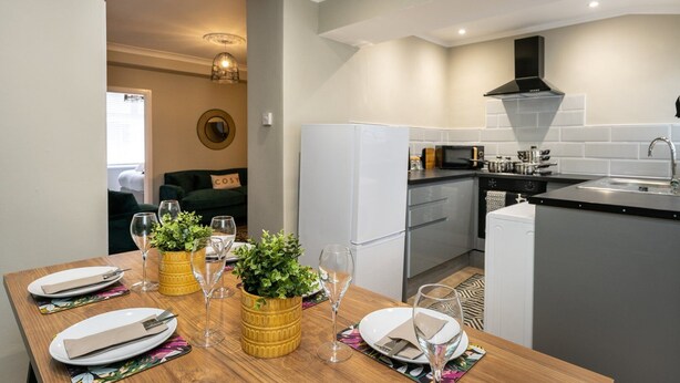 Una cocina moderna con electrodomésticos blancos, una zona de comedor con una mesa de madera preparada para dos personas, y un espacio de estar acogedor con un sofá verde y un letrero que dice 'COSY'.