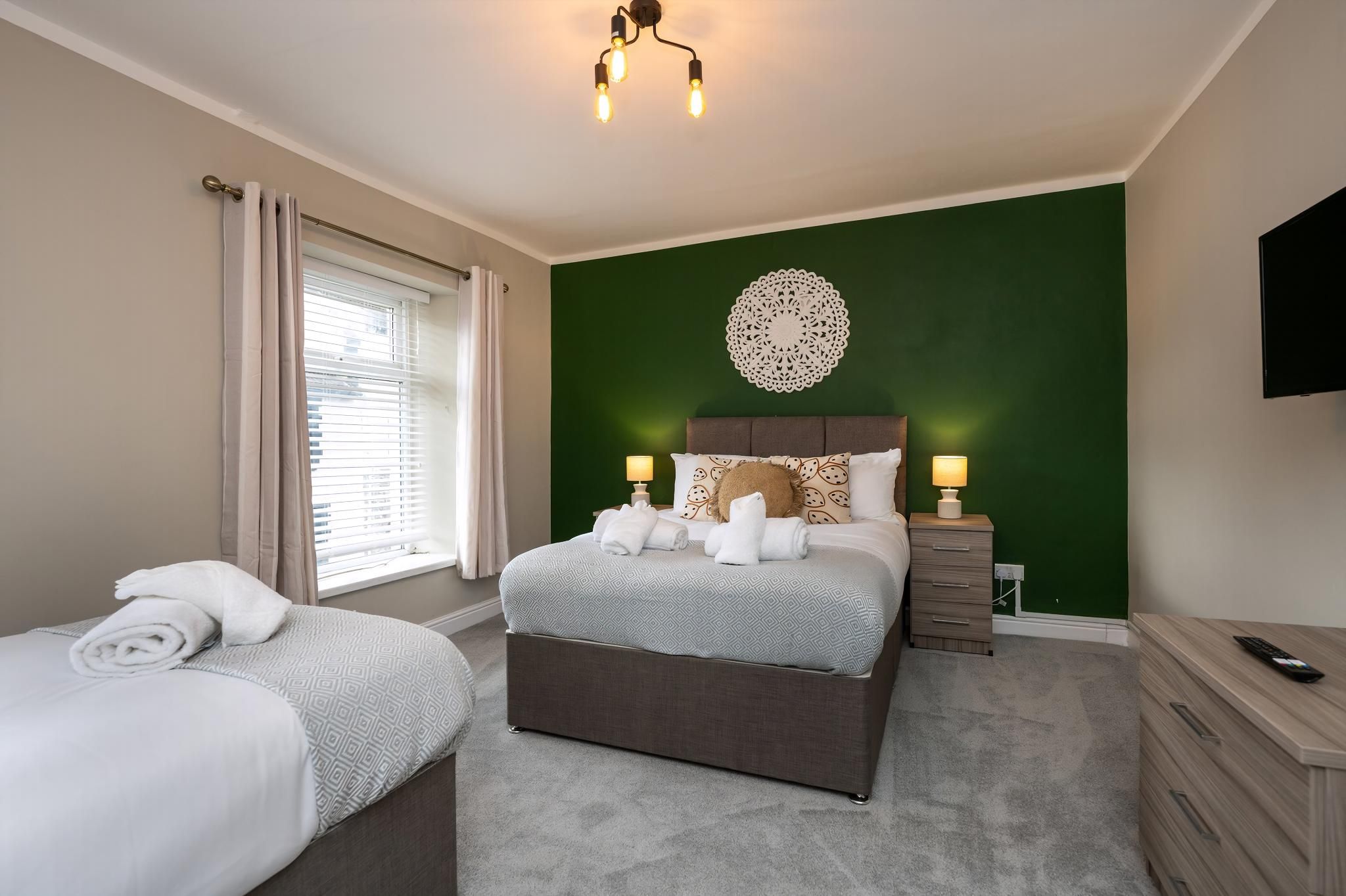 Une chambre d’hôtel comprenant un lit, un petit lit, une table de chevet avec une lampe, une commode et un téléviseur fixé au mur.
