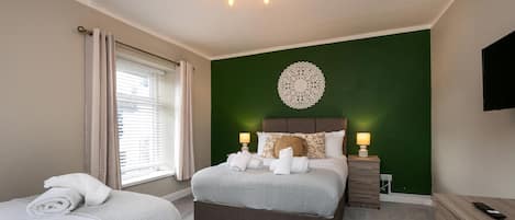 A hotel room with a bed, a smaller bed, a nightstand with a lamp, a dresser, and a TV mounted on the wall.