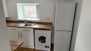 A compact kitchen with a washing machine, a refrigerator, a window with a view, and wooden flooring. - Sea Breeze 1-bed Retreat With Parking (Swansea)