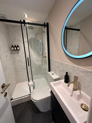 Un baño moderno con ducha envuelta en vidrio, inodoro, lavamanos con grifo dorado y espejo con luz azul.