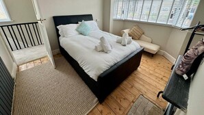 A bedroom with a bed, a window with blinds, a wooden floor, a patterned rug, and a cat on a chair. - Cosy 2 Bedroom Home With Parking Near City Centre (Swansea)