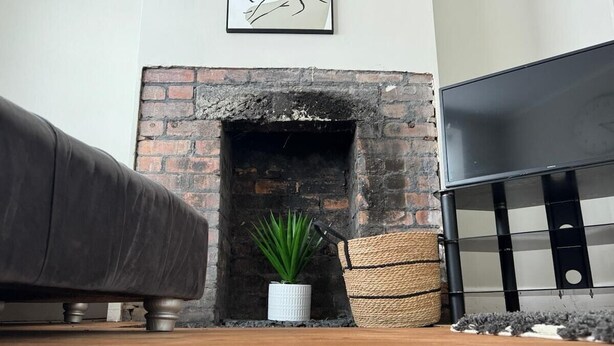A room with a brick fireplace, a white potted plant, a woven basket, and a dark-colored sofa. - Cosy 2 Bedroom Home With Parking Near City Centre (Swansea)