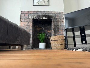 A room with a brick fireplace, a white potted plant, a woven basket, and a dark-colored sofa. - Cosy 2 Bedroom Home With Parking Near City Centre (Swansea)