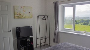 A bedroom with a bed, a TV on a stand, a window with curtains, and a painting on the wall. - Swansea 3 Bedroom Home With Parking & Garden (Swansea)