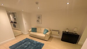 A living room with a beige sofa, a blue rug, a black cabinet, and a painting on the wall. - City Escape 1 Bedroom Flat in Swansea Centre (Swansea)