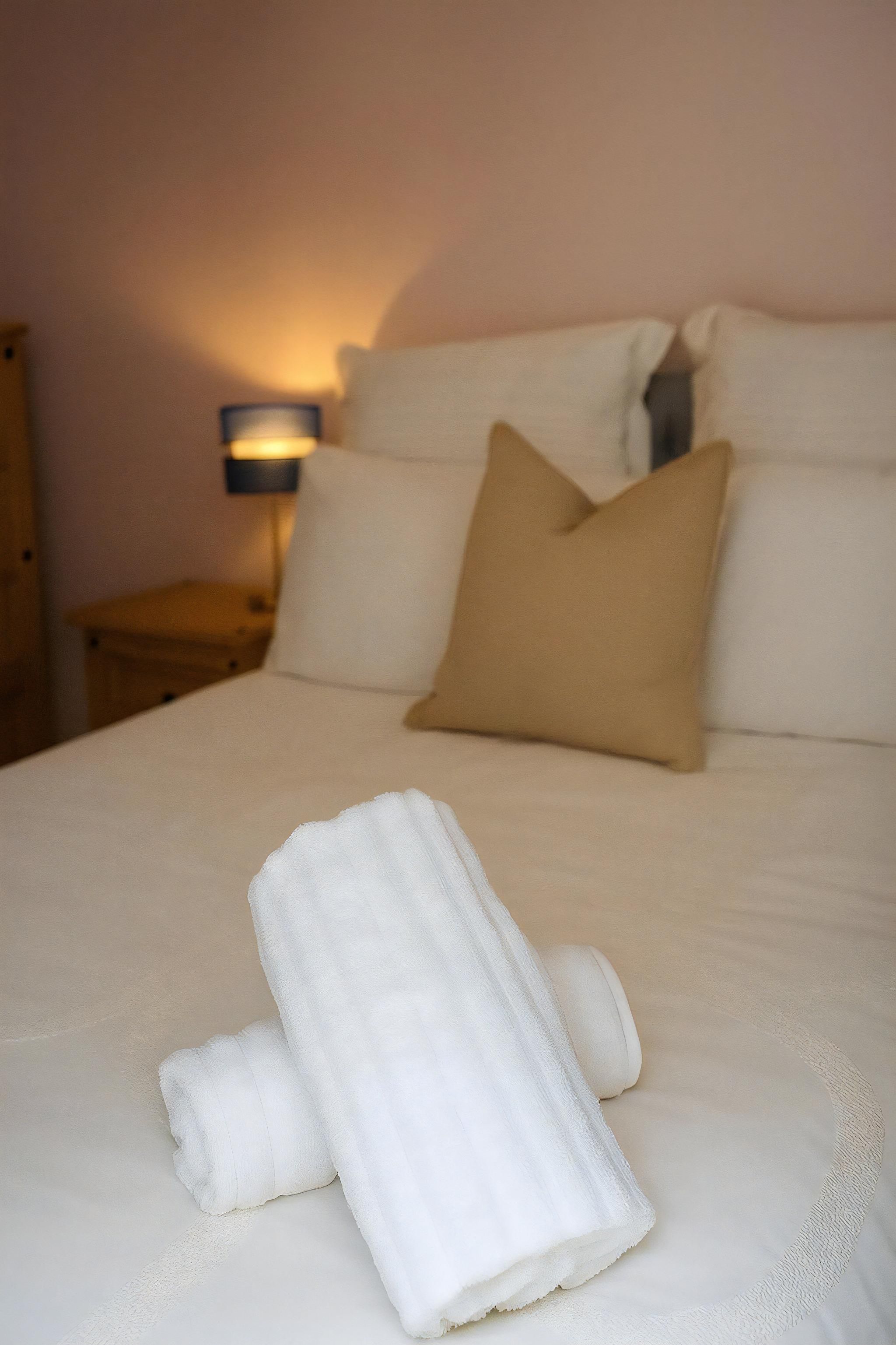 A neatly made bed with white linens and a beige pillow, a bedside lamp, and a wooden nightstand.