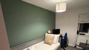 A single bed with a white mattress and beige pillows, a desk with a chair, a radiator, and a patterned ceiling light fixture. - Reading Retreat 1 Bed With Private Parking (Reading)