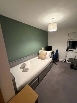 A single bed with a white mattress and beige pillows, a desk with a chair, a radiator, and a patterned ceiling light fixture. - Reading Retreat 1 Bed With Private Parking (Reading)