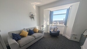 Un salon avec un canapé gris, un petit fauteuil et une grande fenêtre donnant sur des immeubles.