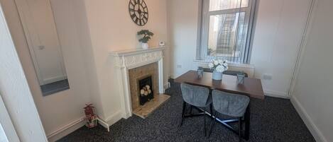 A room with a fireplace, a dining table with two chairs, a clock on the wall, and a window with blinds.