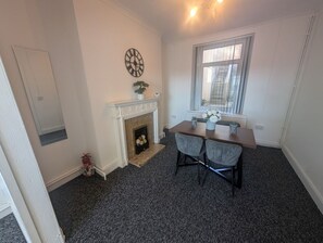 A room with a fireplace, a dining table with two chairs, a clock on the wall, and a window with blinds. - Comfortable 3 Bedroom House With Easy City Access (Swansea)