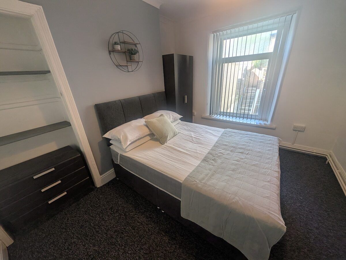 A bedroom with a bed, a wardrobe, a built-in desk, a window with blinds, and a wall-mounted shelf.