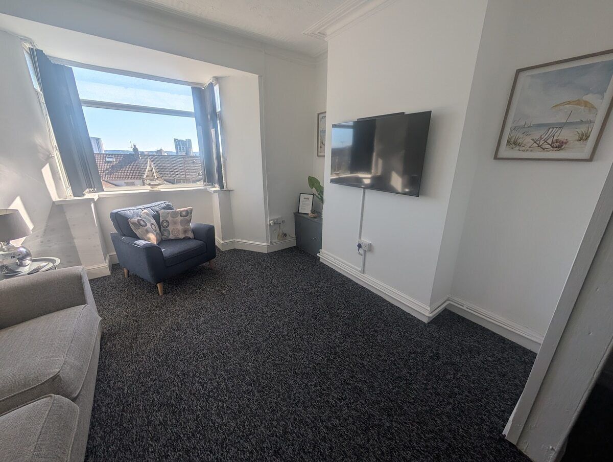 Ein Wohnzimmer mit grauem Teppich, einer grauen Couch, einem blauen Sessel, einem an der Wand montierten Fernseher und einem Fenster mit Blick auf Gebäude.