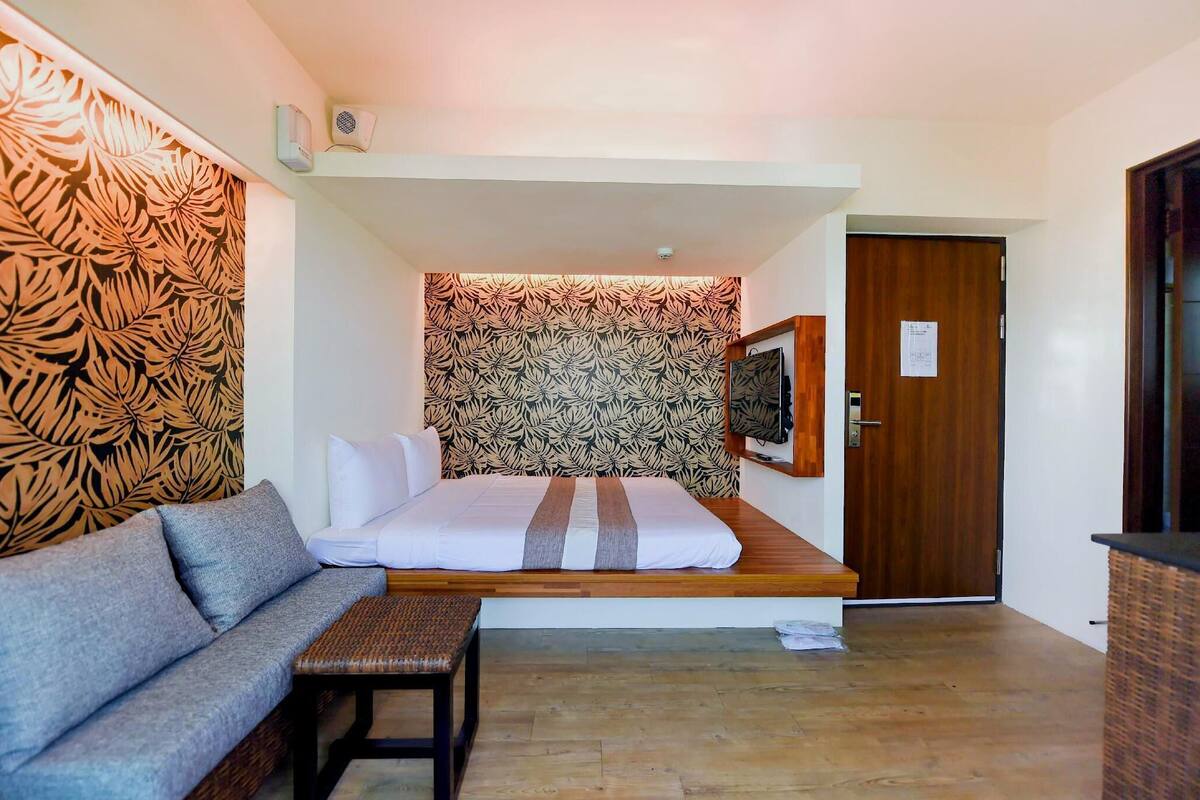 A compact room with a bed, a sofa, a small table, a wall-mounted TV, and a door.