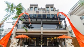 Entrada de hotel com design moderno, fachada de vidro, palmeiras e detalhes decorativos em laranja.