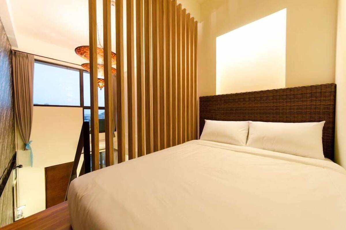 A hotel room with a large bed, a view through a window, and a vertical wooden panel.
