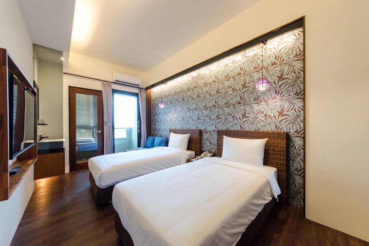 A hotel room with two beds, a TV, a desk, and a window with blinds.