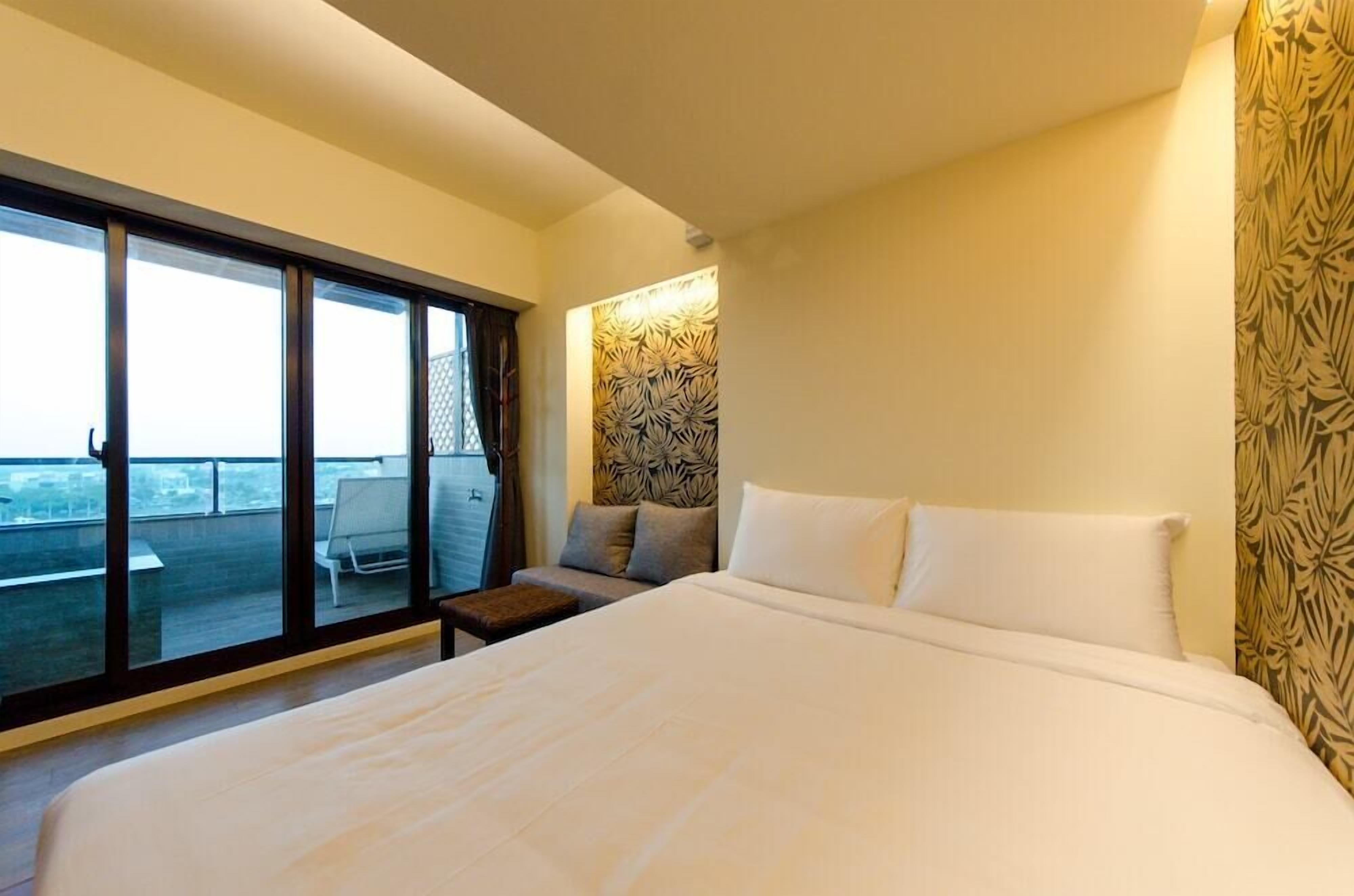 A hotel room with a large bed, a sofa, a small table, and a view through a sliding door to an outdoor balcony.