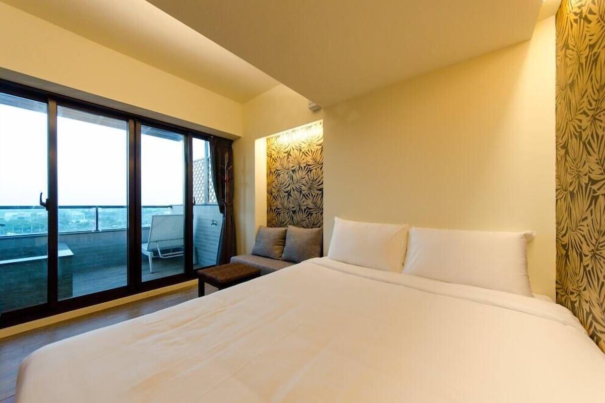 A hotel room with a large bed, a sofa, a small table, and a view through a sliding door to an outdoor balcony.