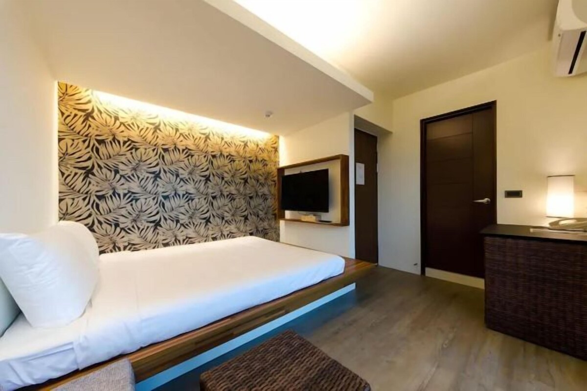 A modern hotel room with a bed, a flat-screen TV, a wooden headboard, and a small table with a lamp.