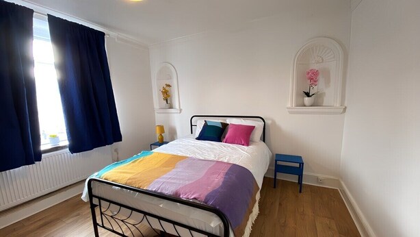 A bedroom with a bed, a nightstand, a window with curtains, and a wall-mounted shelf with a flower.
