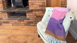 A room with a brick fireplace, a white armchair with a purple and white throw blanket, and a wooden floor.