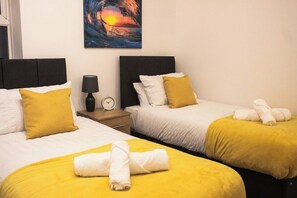 A hotel room with two beds, each with a yellow bedspread and white pillows. There is a painting of a sunset on the wall, a clock on the nightstand, and towels on the beds. - Family-friendly 3-bed House in Swansea (Swansea)
