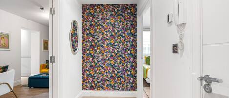 A hallway with a floral-patterned wall, a door with a phone, and a glimpse of a room with a bed and a yellow chair.