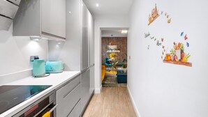 A modern kitchen with white cabinetry, a built-in oven, and a countertop with two appliances. A hallway leads to a dining area with a colorful brick wall and a chandelier. - Brighton Seaside 1-bed Retreat Near The Lanes (Brighton)