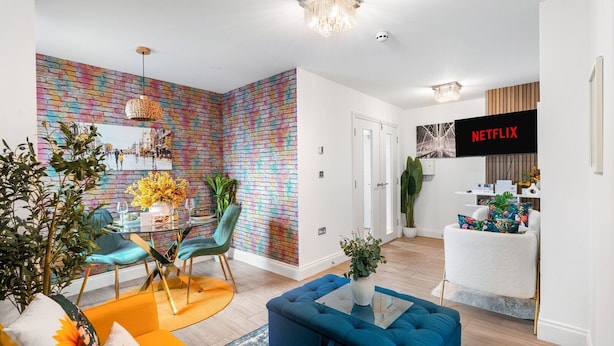 A modern living room with a colorful brick wall, a blue sofa, a dining area with a glass table, and a kitchen area with a Netflix sign. - Brighton Seaside 1-bed Retreat Near The Lanes (Brighton)