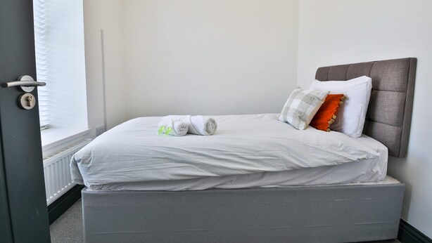 Una cama bien tendida con sábanas blancas y cabecera gris, una ventana con persianas, y una puerta con pomo.