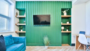 Una sala de estar moderna con una pared verde de acento, un televisor de pantalla plana, un sofá azul, piso de madera y un escritorio con una silla.