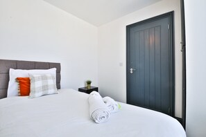 A neatly made bed with white linens and rolled towels, a grey upholstered headboard, and a dark wooden door. - Bright 3-bed Swansea House + Parking (Swansea)