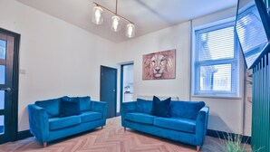 Un salón amplio con dos sofás azules, suelo de madera con diseño espiga, lámpara moderna, ventana grande con persianas y un cuadro en la pared.