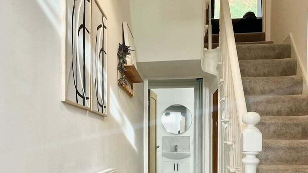 Un couloir avec un radiateur, un pouf bleu, un escalier avec des balustrades blanches et une vue sur une salle de bain avec un miroir et un lavabo.