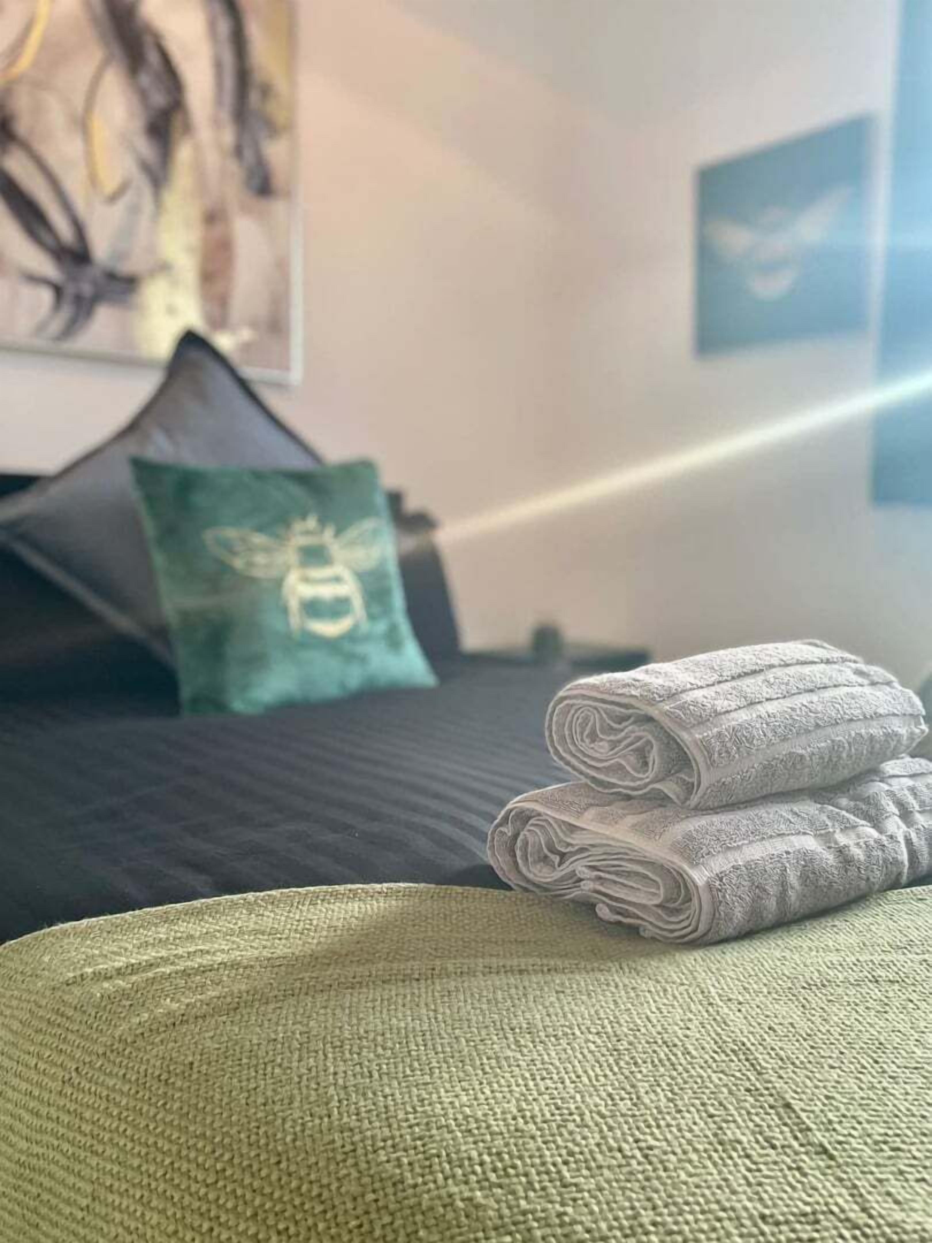 A neatly made bed with rolled towels, a green pillow with a bee design, and a dark-colored headboard.