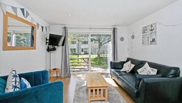 A living room with a blue sofa, a black sofa, a wooden coffee table, a TV mounted on the wall, and a large sliding glass door leading to an outdoor area.